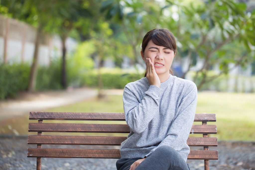 How to Prevent Cavities and Protect Your Oral Health Cavities 1 A woman sitting on a bench outdoors holding her cheek in pain, showing discomfort from a cavity.