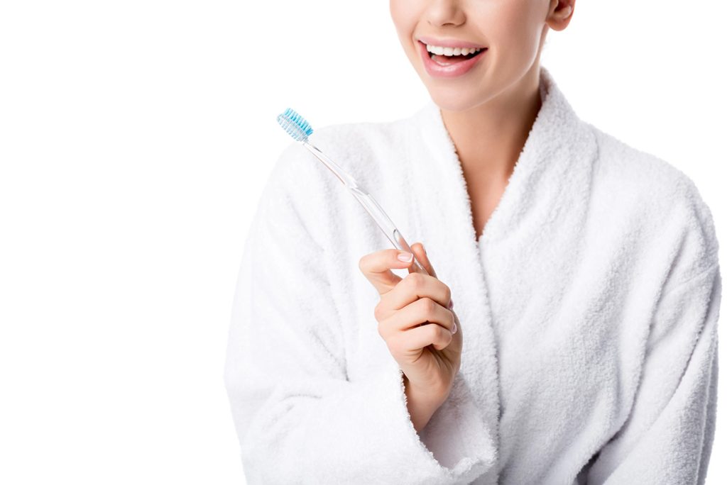 How to Properly Brush & Floss 1 A woman in a white bathrobe smiling while holding a toothbrush with blue bristles against a plain white background.